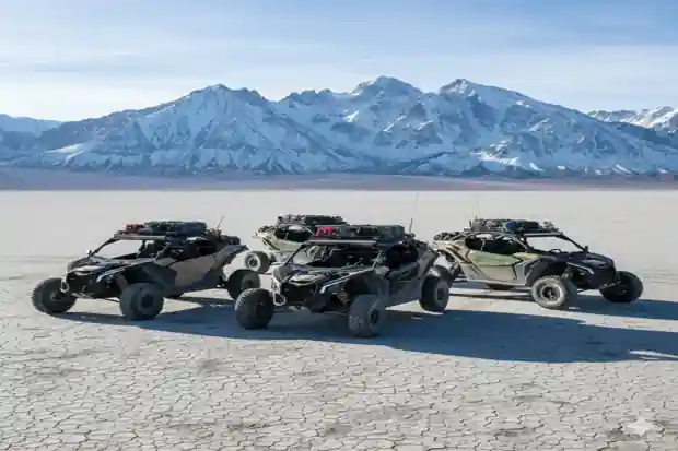 Three off-road dune buggies parked on a dry salt flat with snow-capped mountains in the background